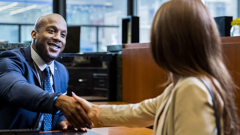 Male bank worker in navy blue suit, smiling and leaning over desk to shake hands with female bank customer in financial office.